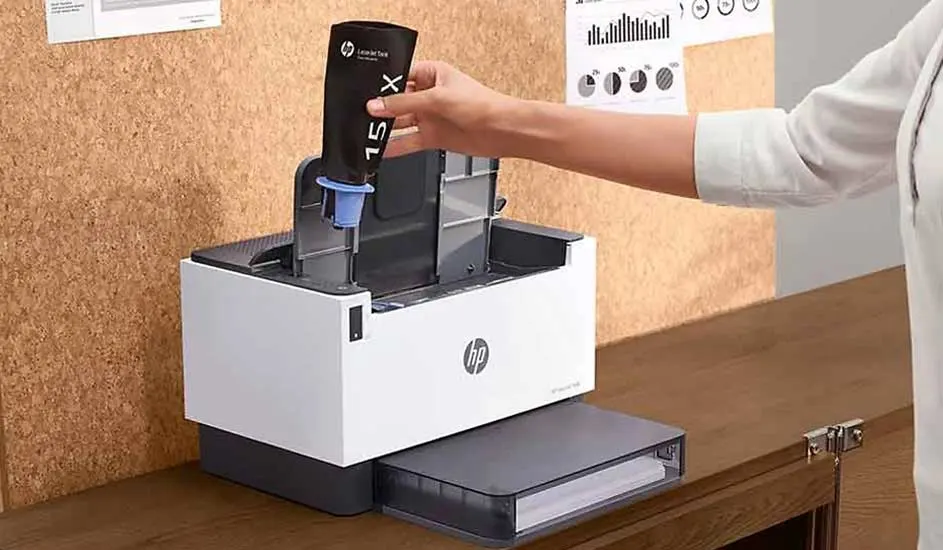 Een persoon plaatst een zwarte HP 15X tonercartridge in een witte HP laserprinter op een houten bureau.