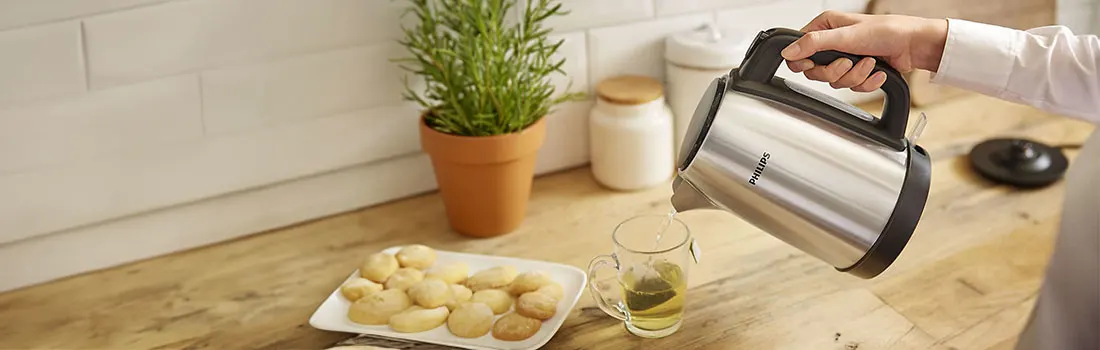 Vue de la cuisine avec une bouilloire en inox versant de l'eau chaude dans une tasse de thé, des biscuits et une plante.