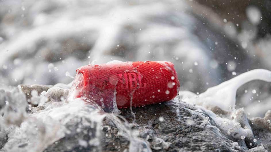 Een rode JBL-luidspreker ligt op een rots, omringd door opspattend water. De achtergrond is wazig door de beweging van het water.