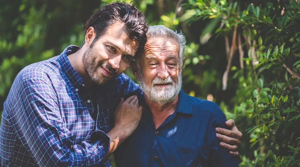 Outdoor shot with green foliage background. One wearing a blue and white checkered shirt, the other is wearing a blue button-up shirt.