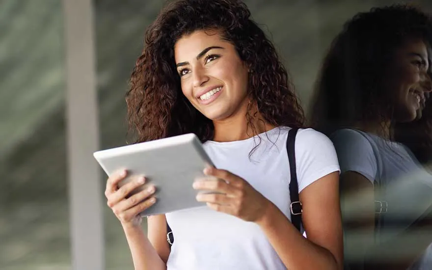 Une femme souriante aux cheveux bouclés tient une tablette, son reflet apparaît dans la vitre à côté d’elle.