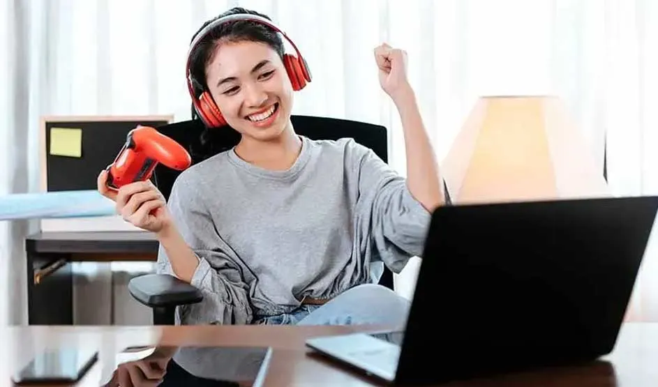 A young woman in red headphones smiles and cheers, holding a red game controller while playing on a laptop at the table.