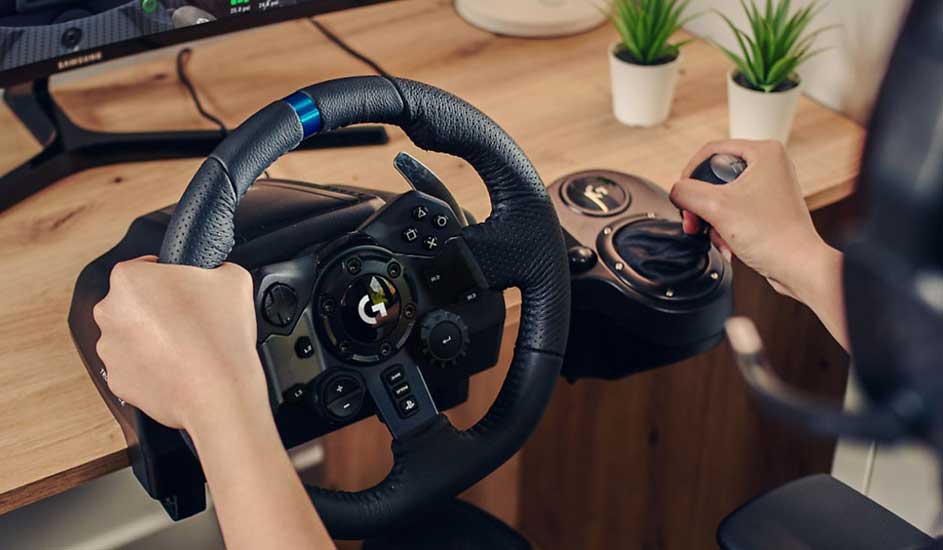 Een zwarte racestuur en versnellingspook op een houten bureau, met een monitor en planten op de achtergrond.