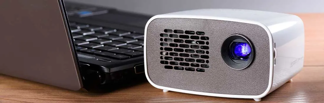 Een compacte projector staat naast een laptop op een houten bureau, klaar om video of presentaties helder weer te geven.