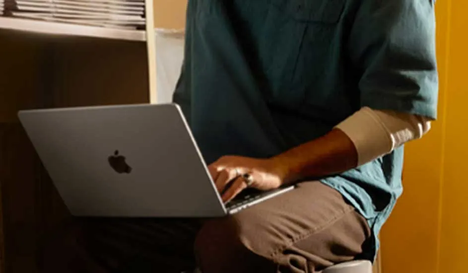 Een laptop met een zilveren behuizing wordt gebruikt in een kamer met een gele muur en een boekenkast.
