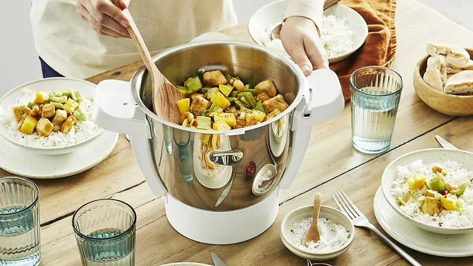 Houten tafel met een zilverkleurige kookpot, gevuld met groenten en blokjes vlees, naast borden met rijst en glazen water.