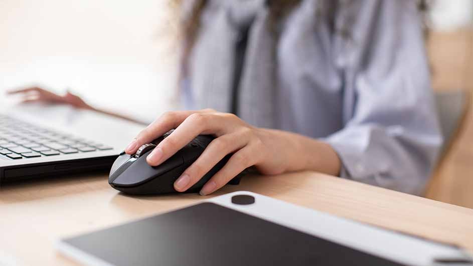 Une main sur une souris noire près d'un ordinateur portable et d'une tablette sur un bureau en bois clair.