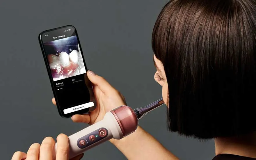 Une femme utilise la brosse à dents tout en regardant ses dents en direct sur son smartphone. Le manche possède plusieurs boutons.