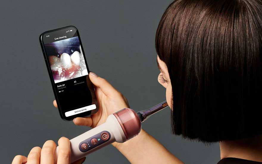 Een vrouw gebruikt de tandenborstel terwijl ze op haar smartphone een livebeeld van haar tanden bekijkt. De borstel heeft meerdere knoppen.