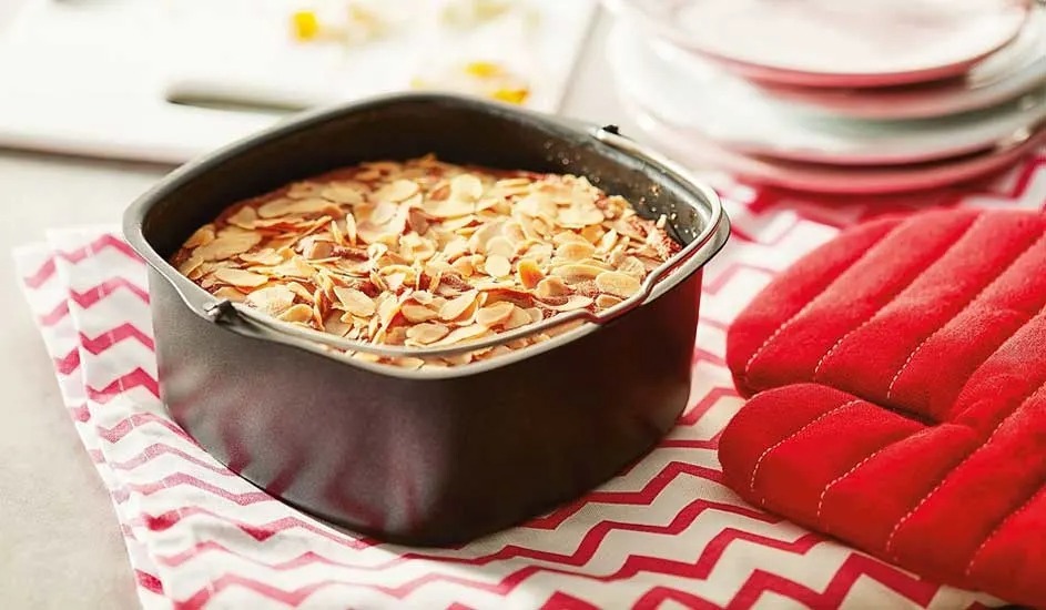 Flaked almonds cover a golden baked cake in a black tin, beside a red oven glove on a patterned cloth.
