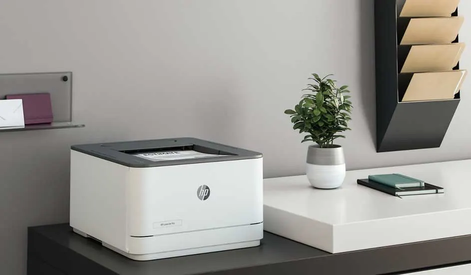 Een wit HP-printer staat op een donker bureau. Een plant en notitieboekje staan op een wit bureau ernaast.