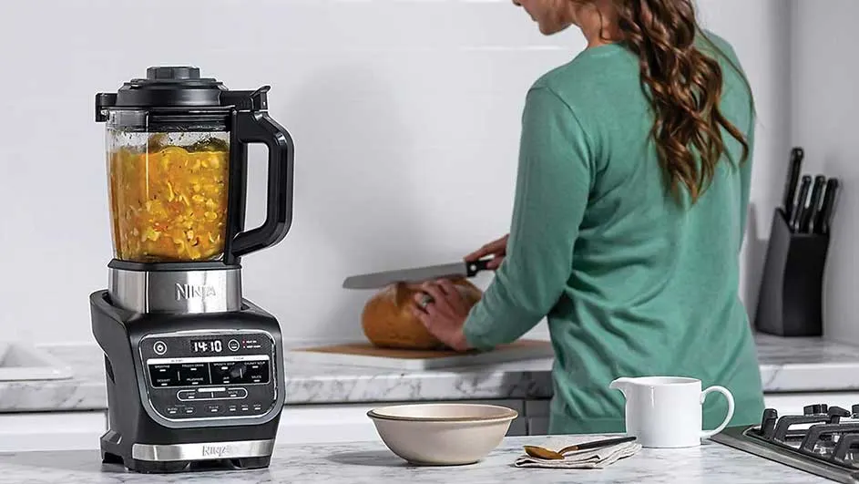 Un mixeur noir et argenté rempli de soupe orange, à côté d'une courge coupée sur un comptoir de cuisine clair.