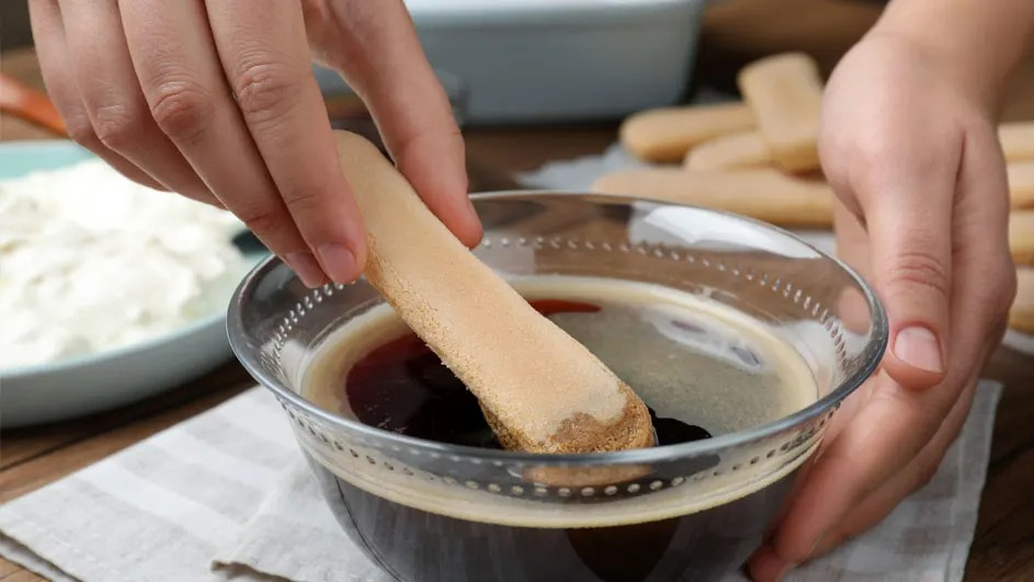 Un biscuit trempé dans un bol en verre rempli de café sur une table en bois. D'autres biscuits et un plat de crème à l'arrière-plan.