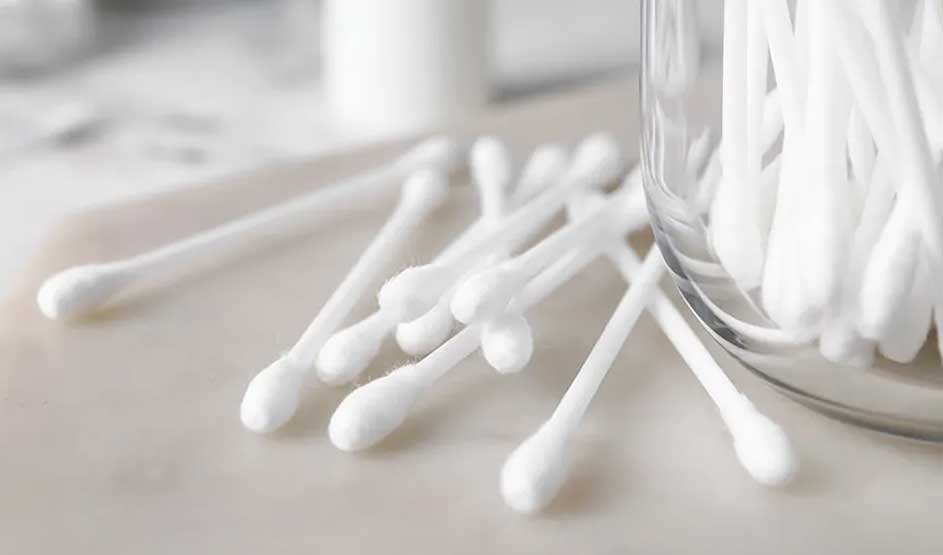 Des cotons-tiges blancs sont dispersés sur une surface claire, à côté d’un pot en verre transparent rempli d’autres bâtonnets assortis.