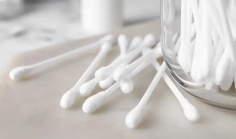 Des cotons-tiges blancs sont dispersés sur une surface claire, à côté d’un pot en verre transparent rempli d’autres bâtonnets assortis.