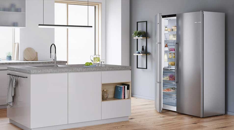 Cuisine moderne avec îlot blanc, évier, et grand frigo en inox à porte ouverte, sur parquet clair. Étagère murale visible.