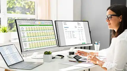 Une table blanche avec un ordinateur portable, deux écrans affichant des feuilles de calcul, une calculatrice et des fournitures de bureau.