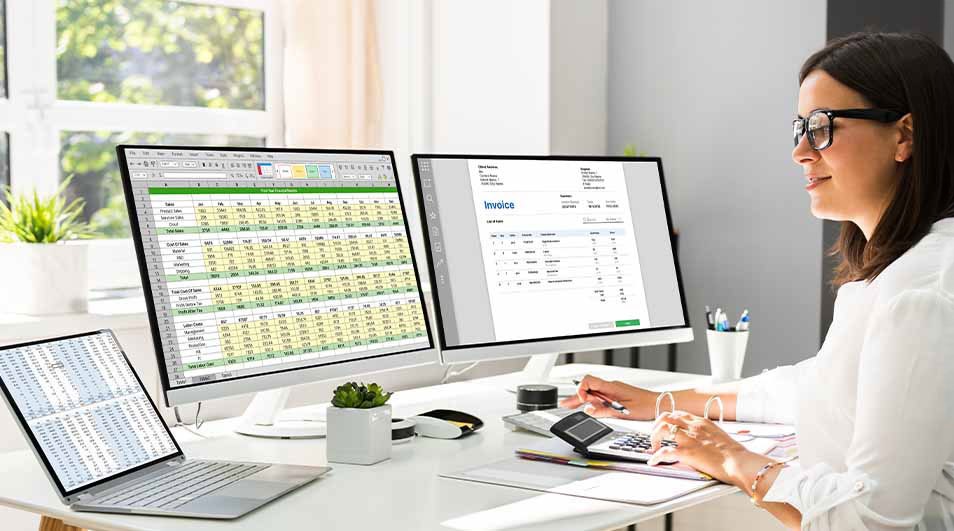 A desk with a laptop, two monitors displaying spreadsheets and invoices, a calculator, and office supplies.