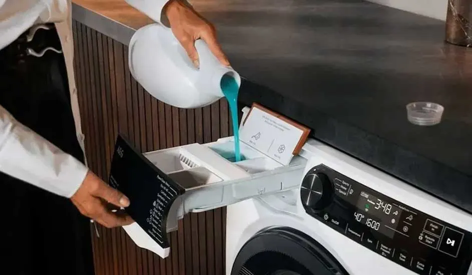 Een persoon giet vloeibaar wasmiddel in het doseerbakje van een AEG-wasmachine met digitaal display en houten zijpaneel.