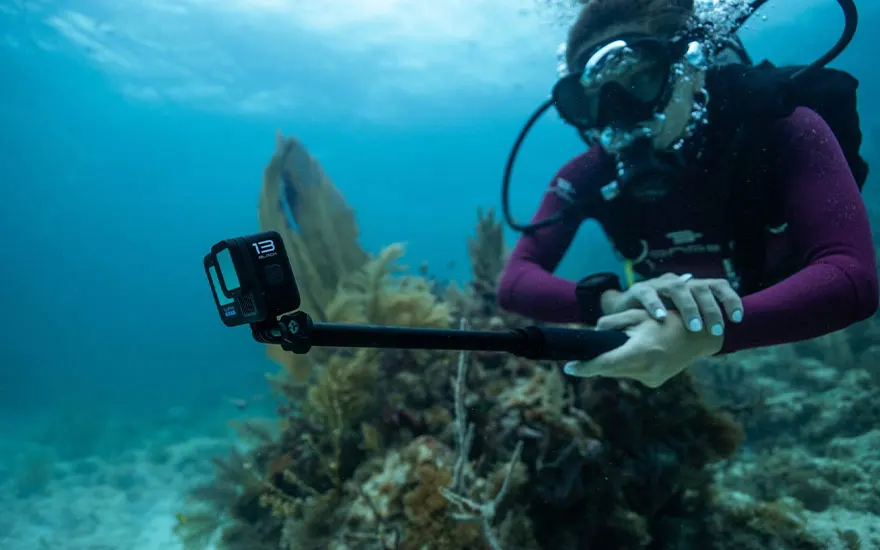 Een duiker met duikuitrusting houdt een GoPro vast onder water, met koraal en blauw water op de achtergrond.