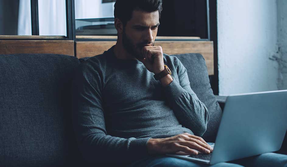 Un ordinateur portable est posé sur un canapé gris. Une personne porte un pull gris et une montre. Un mur blanc est visible à l'arrière-plan.