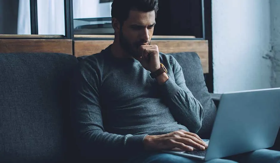 Een persoon zit op een grijze bank met een laptop. De achtergrond is een mix van houten panelen en een witte muur.