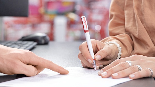 Une main signe un document blanc avec un stylo rouge et blanc. Une autre main pointe le papier. À l'arrière, un clavier et un écran.