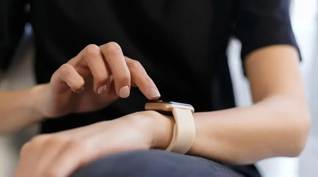 Een hand tikt op een smartwatch met een beige band, gedragen aan de pols. De achtergrond is wazig.