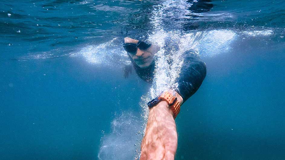 Un nageur en lunettes nage dans l'eau bleue. Une montre-bracelet est visible. Des bulles montent vers la surface.