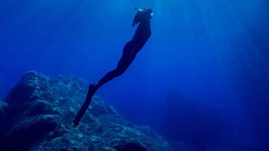 Un plongeur en combinaison noire descend dans l'eau bleue, près d'un rocher. La lumière perce depuis la surface.