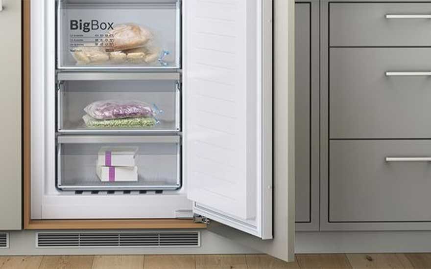 Un congélateur blanc ouvert révèle trois tiroirs avec des aliments congelés, à côté d'armoires grises.