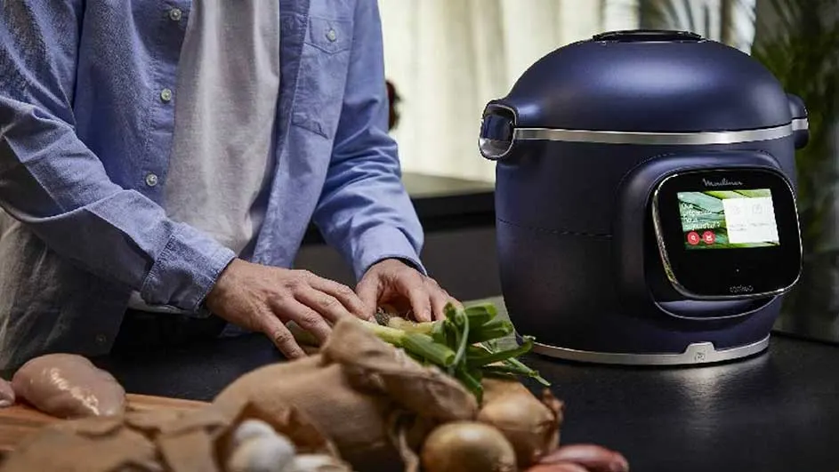 Een blauwe multicooker naast groenten en kip op een houten plank.