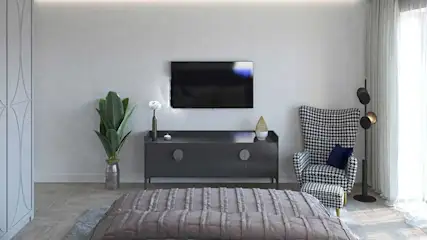 A room with a TV above a gray cabinet, a patterned chair, and a bed in the foreground. Curtains on the right.