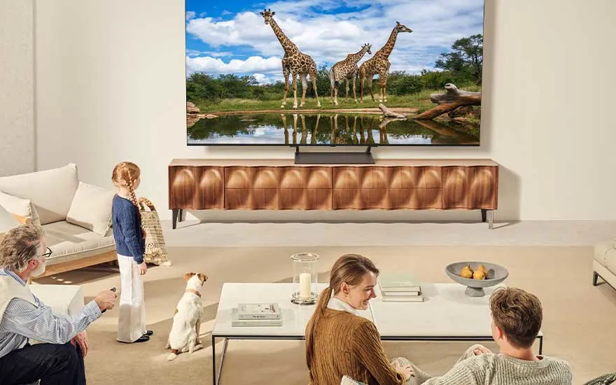 Een gezin en hond kijken in een lichte woonkamer naar een LG-tv waarop giraffen bij een plas water te zien zijn, iedereen lijkt ontspannen.