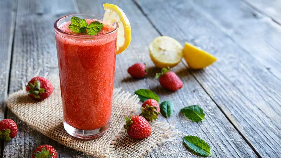 Een glas rode smoothie met citroen en munt staat op een houten tafel, omringd door aardbeien en citroenpartjes.