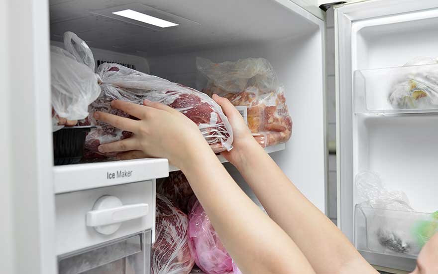 Un congélateur blanc ouvert montre des sacs de nourriture. Une personne tient un sac de viande rouge. Il y a un bac à glaçons à gauche.