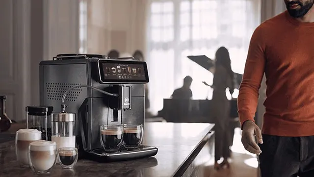 Machine à café noire avec tasses sur un comptoir. Arrière-plan flou avec des personnes et un piano.