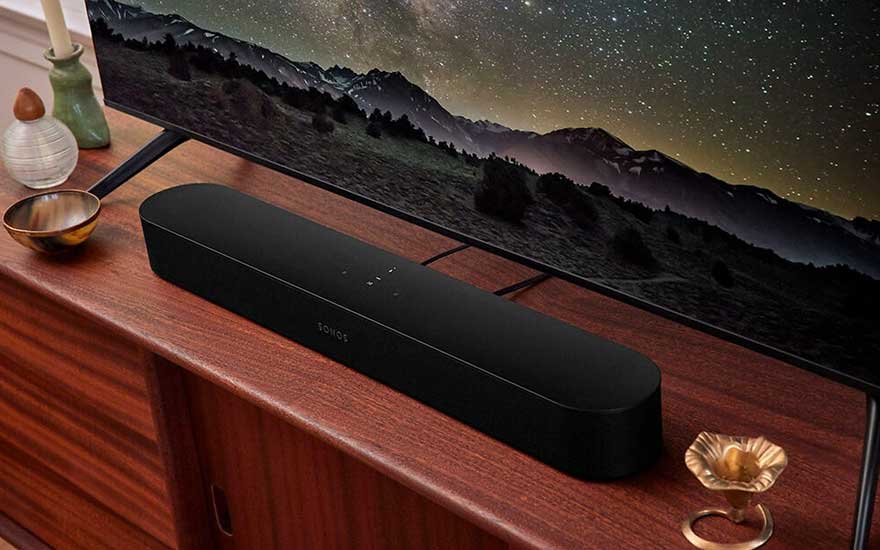 Een zwarte Sonos soundbar ligt op een houten dressoir onder een tv-scherm met berglandschap.