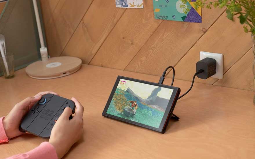 On a desk, a tablet is connected to a charger, displaying a game. A black controller is held in someone's hands. The background shows wooden walls.