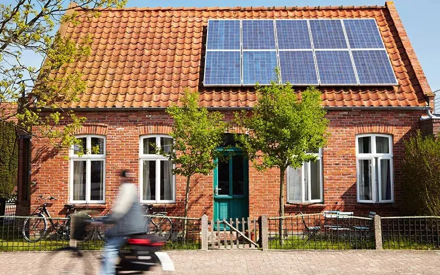 Red brick house with orange tiled roof and solar panels. Green door, white windows, and a fence.