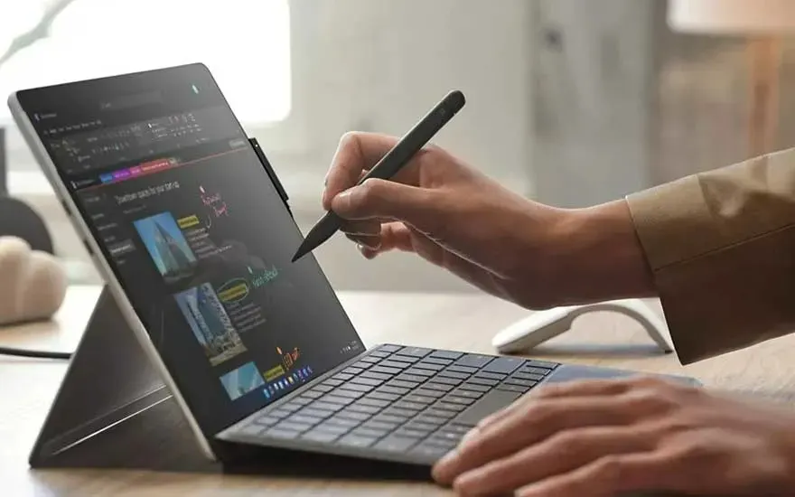 Un ordinateur portable gris est utilisé avec un stylet sur un bureau, avec une souris blanche visible en arrière-plan.