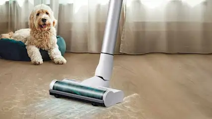 Un aspirateur blanc et argenté sur un tapis brun clair, avec un coussin bleu derrière.