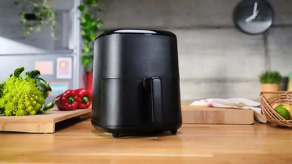 Une friteuse à air noire sur un comptoir en bois, avec des légumes et une horloge en arrière-plan.