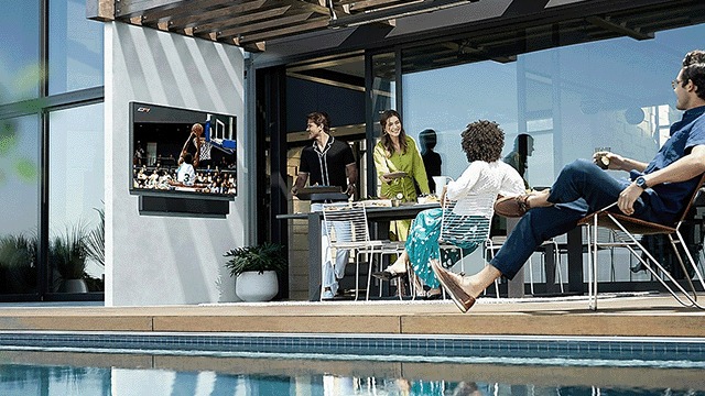Une télévision diffuse un match de basket-ball près d'une piscine turquoise, avec des meubles de patio.