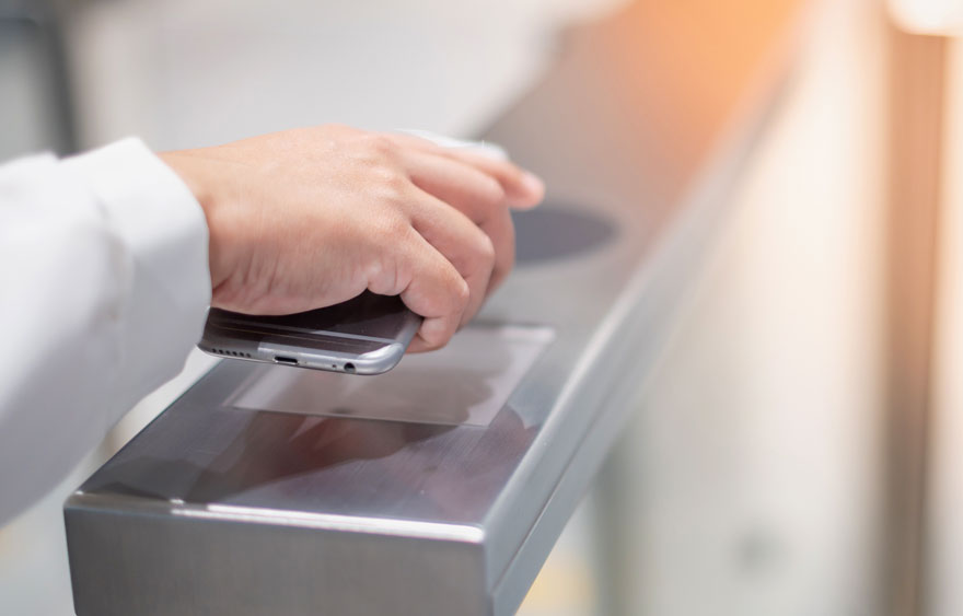 Een hand houdt een zilverkleurige smartphone boven een metalen oppervlak met een transparant vierkant. Op de achtergrond is een witte muur.