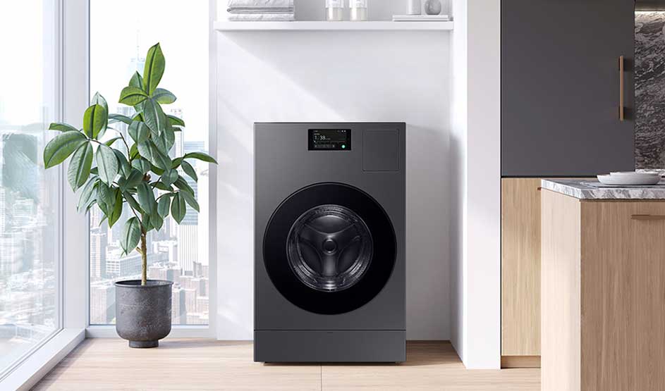 Grijze wasmachine in een nis naast houten keukenfronten; links een grote groene kamerplant bij raam met stadszicht, bovenop witte plank met handdoeken.