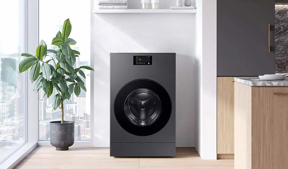 Grijze wasmachine in een nis naast houten keukenfronten; links een grote groene kamerplant bij raam met stadszicht, bovenop witte plank met handdoeken.