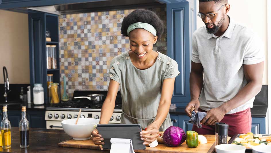 Une femme et un homme dans une cuisine regardent une tablette. Des légumes sont sur une planche à découper.