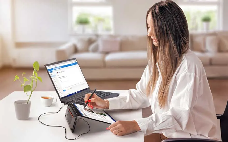 Een vrouw werkt thuis met een laptop en tekent met een stylus op een groot digitaal scherm, naast een plant en een kop koffie.