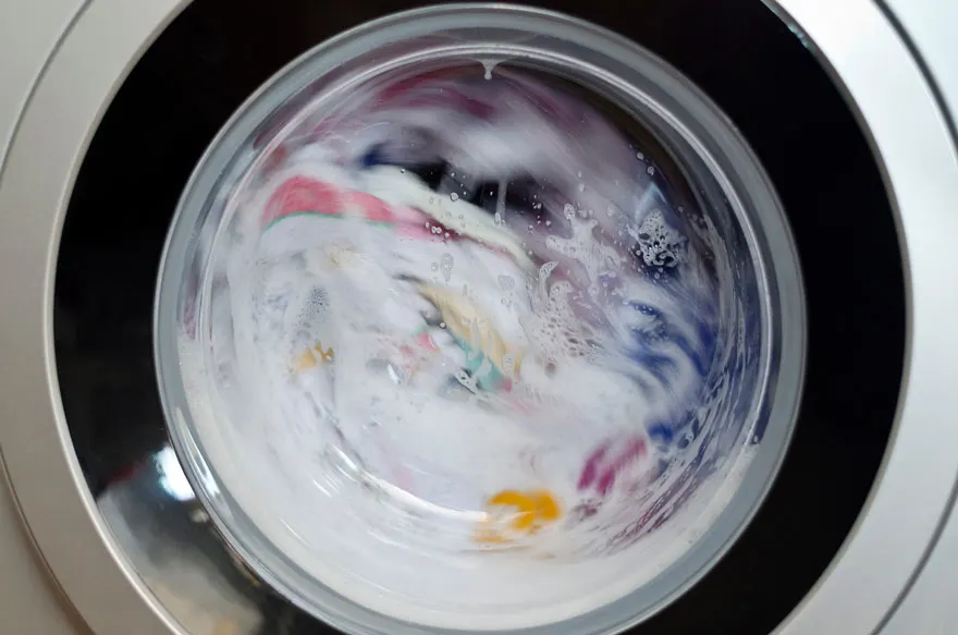 Une machine à laver argentée avec une porte sombre et ronde montre des vêtements colorés en cours de lavage avec de l'eau savonneuse.